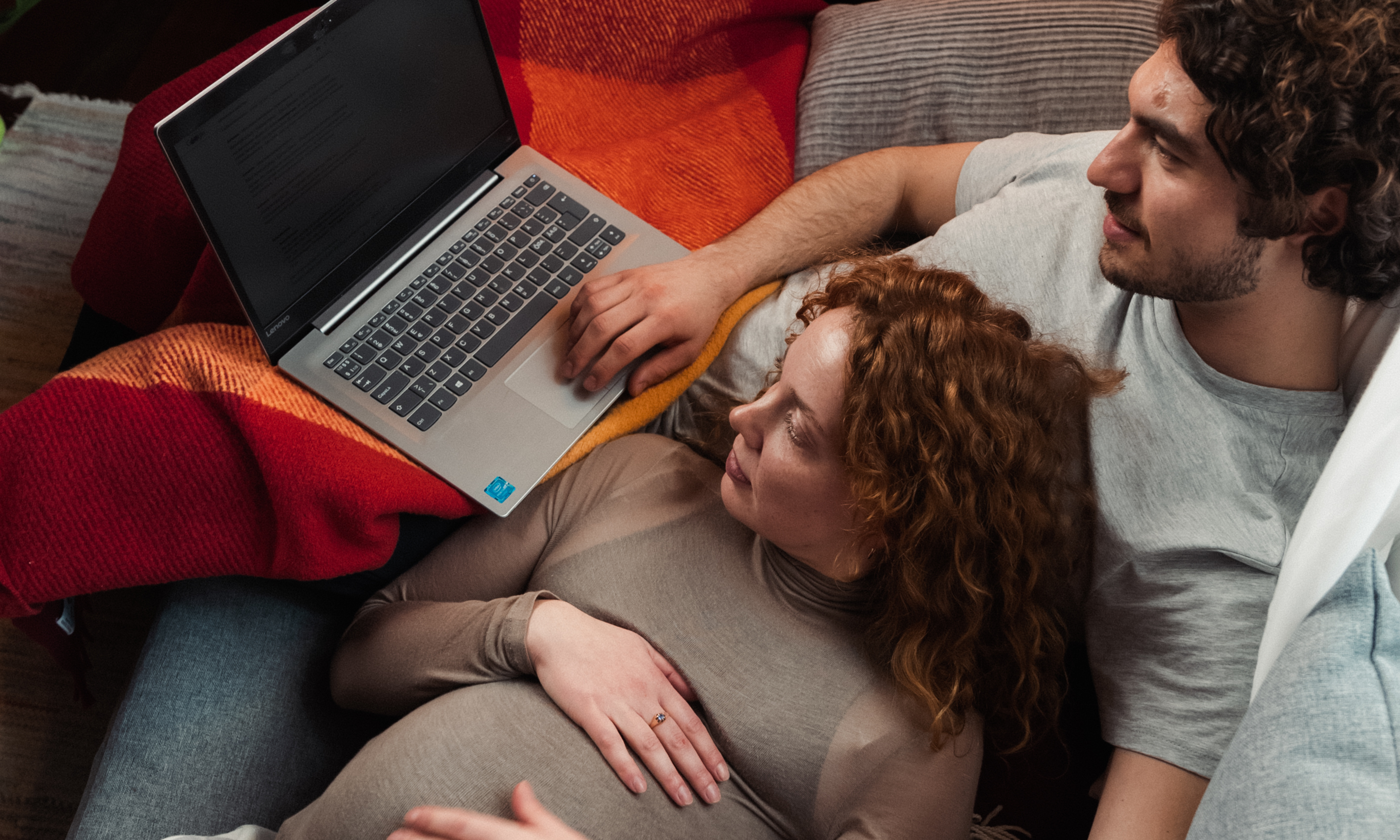 Par, rødhåret gravid kvinde og mørkhåret mand, ligger i sofa og kigger på en computer, fotografi