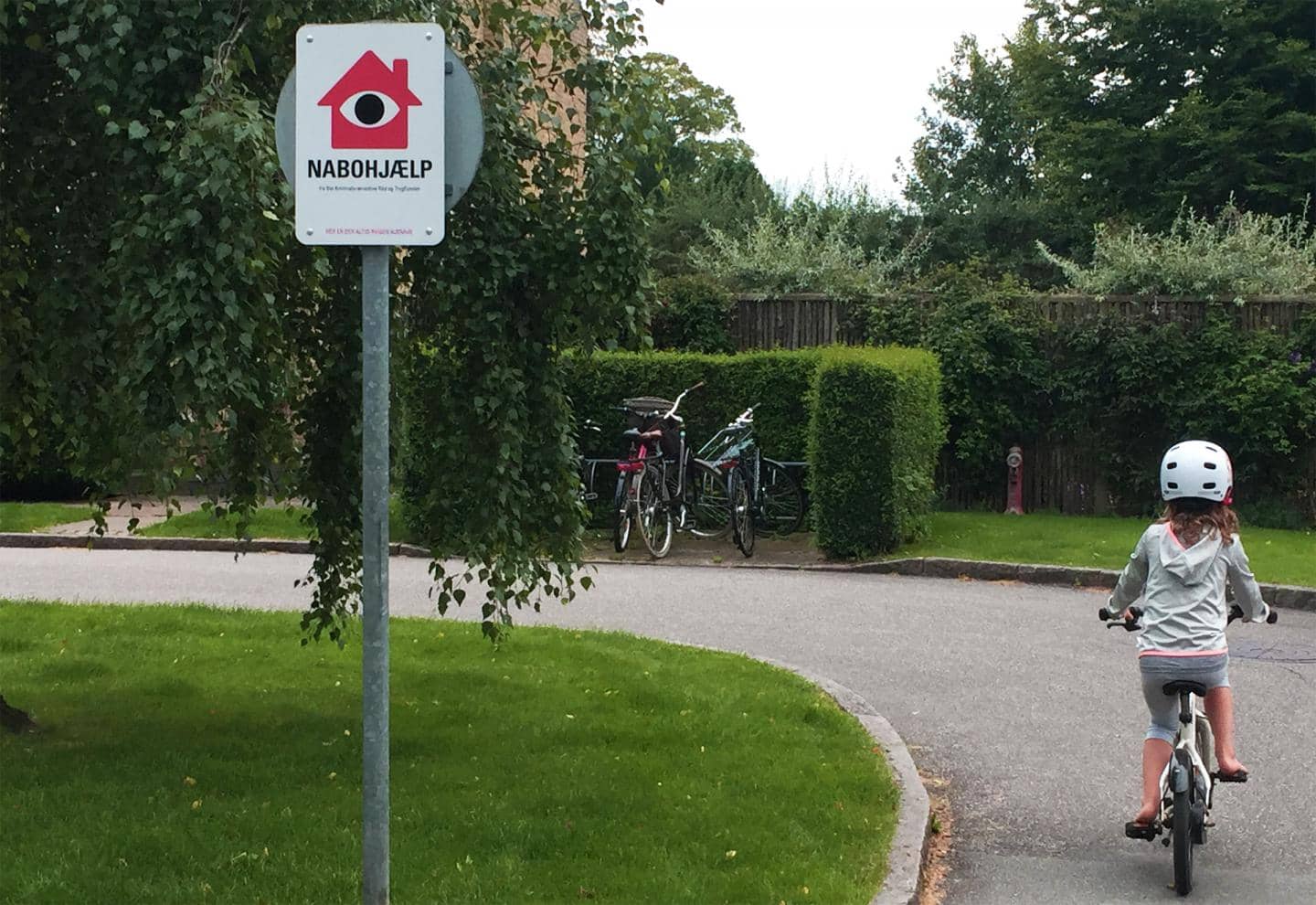 Pige cykler på vej, skilt fra Nabohjælp, fotografi