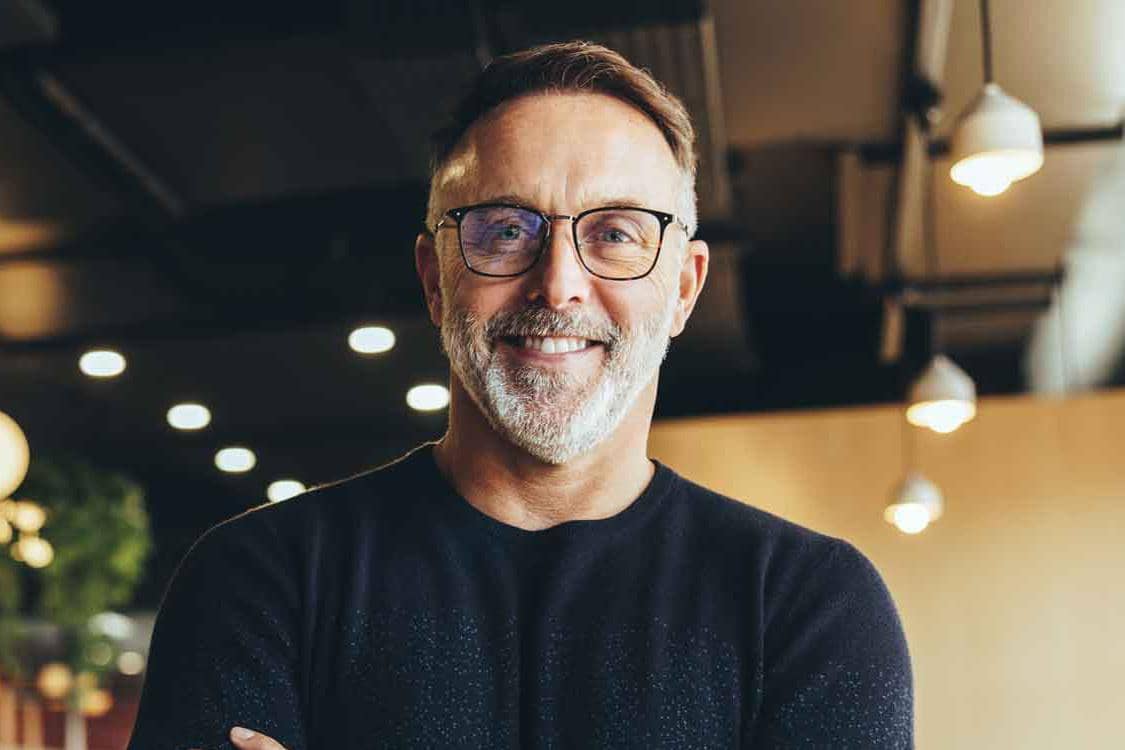 Mand med briller og sort trøje smiler til kameraet, står indendørs under lamper, fotografi